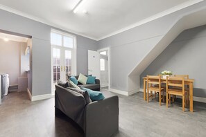 Ein geräumiges Loft mit grau-weißer Farbgestaltung, einem Essbereich mit Holztisch und -stühlen sowie einem grauen Sofa mit Kissen.