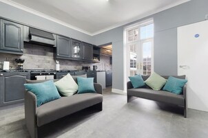 A modern kitchen with grey cabinets and a brick backsplash, two grey sofas with teal cushions, and a large window providing natural light.
