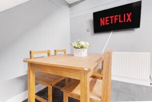 A room with a wooden table, chairs, a wall-mounted TV displaying 'NETFLIX', and a radiator.