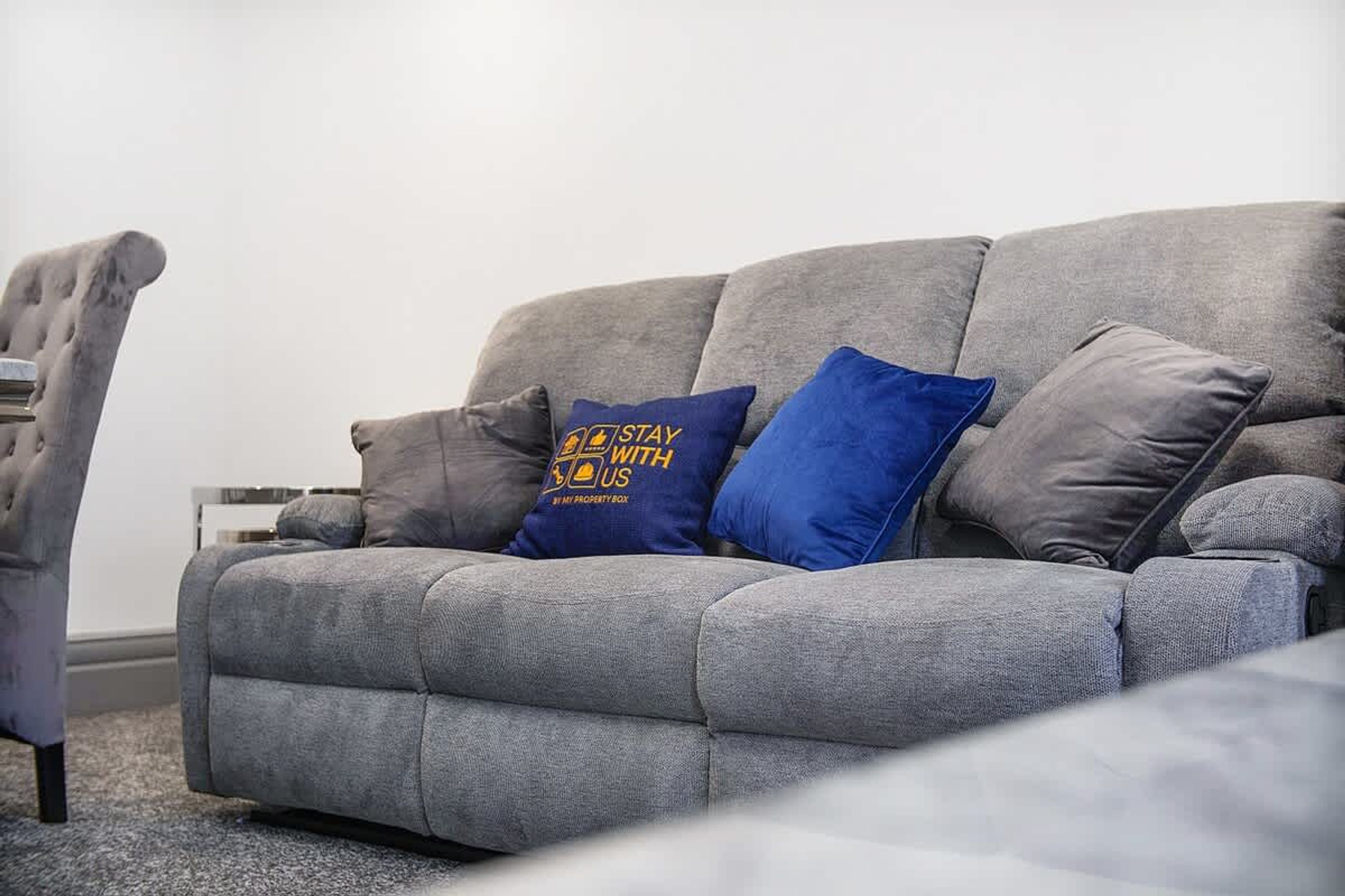 A grey sofa with two pillows, one blue and one grey, in a living room with a plain white wall and a grey carpet.