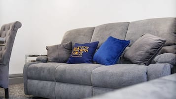 A grey sofa with two pillows, one blue and one grey, in a living room with a plain white wall and a grey carpet.