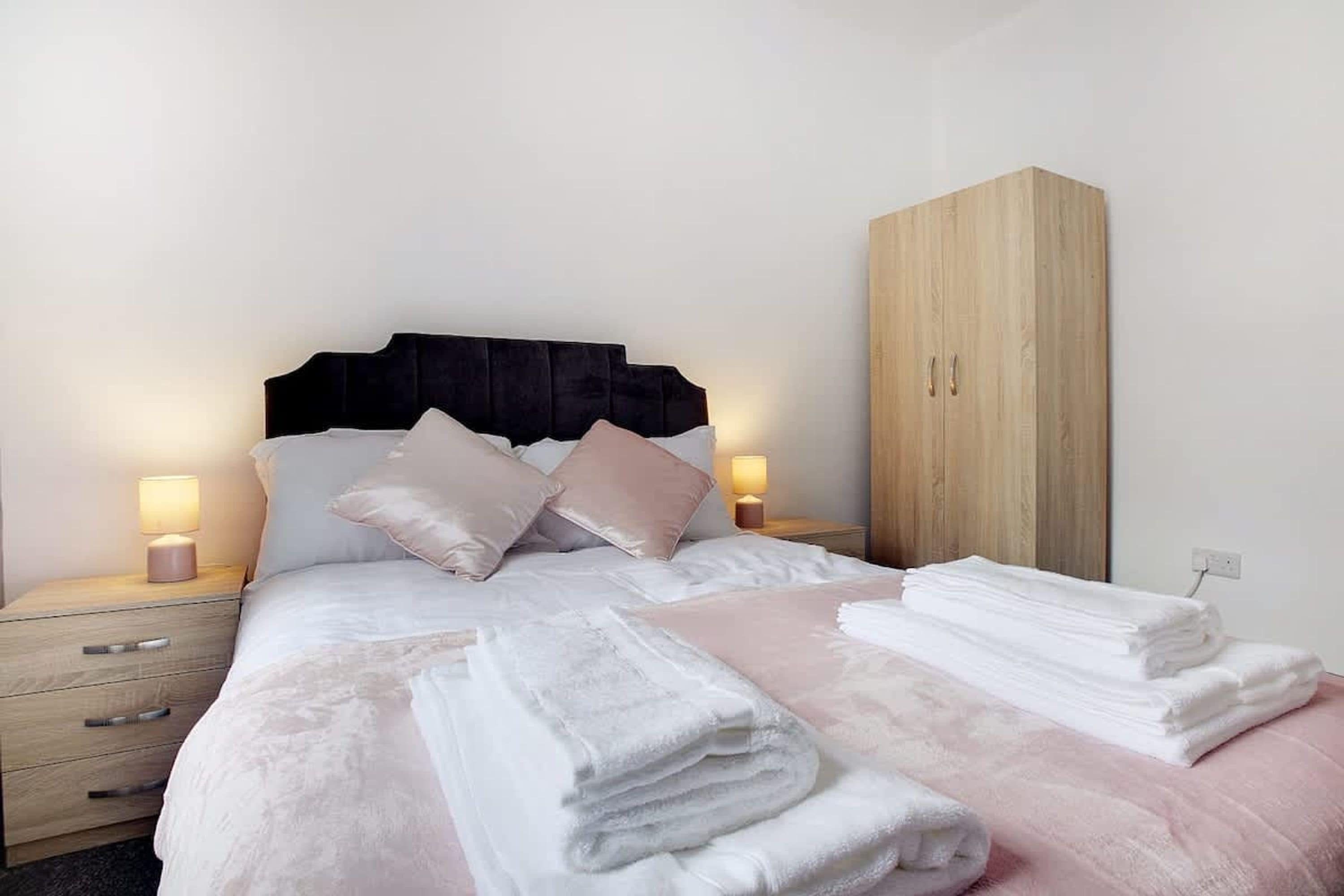 A neatly made bed with pillows and a stack of towels, a wooden wardrobe, and bedside lamps on a nightstand.