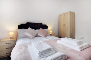 Ein ordentlich bezogenes Bett mit Kissen und einem Stapel Handtüchern, ein Holzschrank und Nachttischlampen auf einem Beistelltisch.