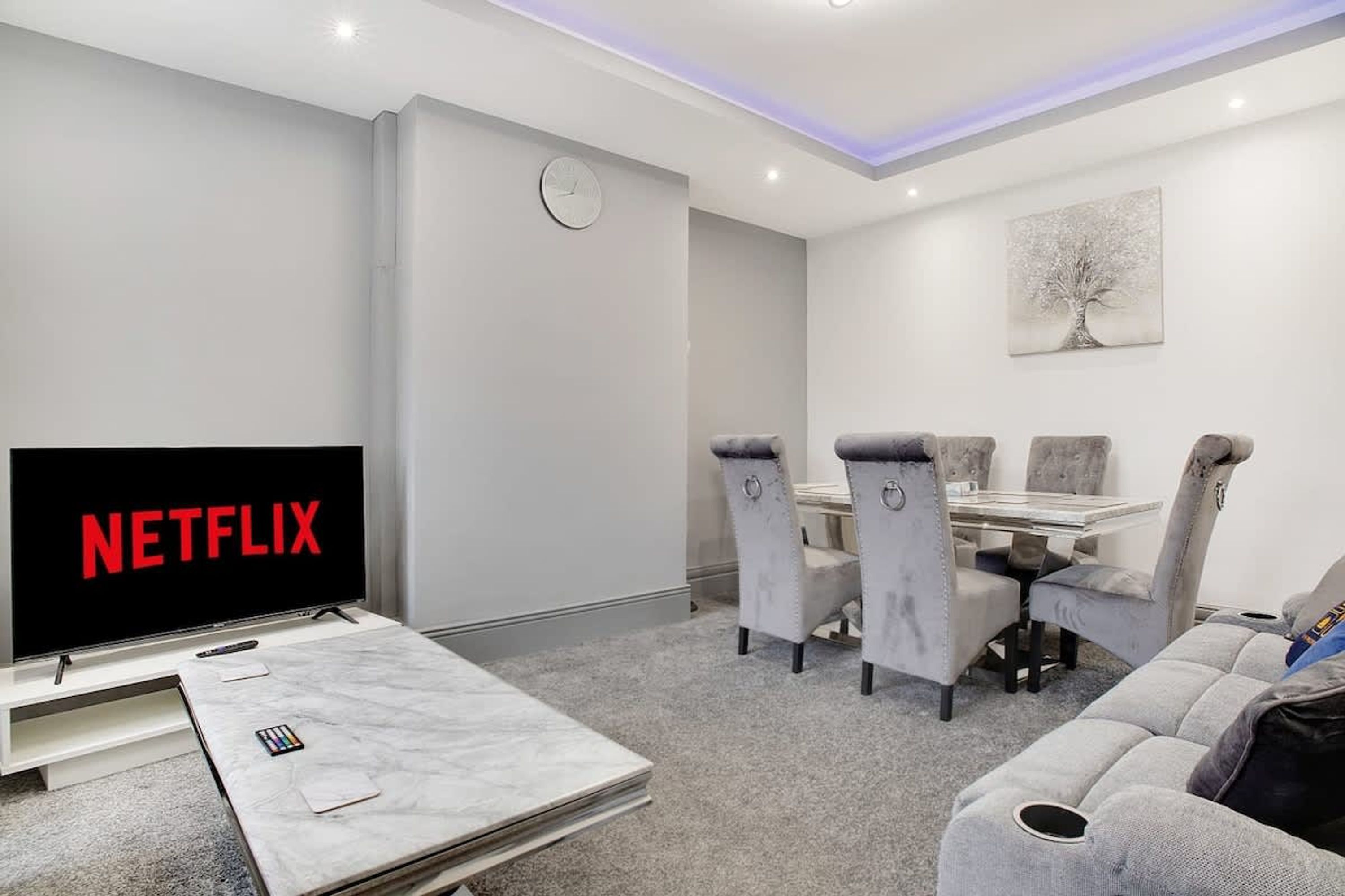 Ein modernes Wohnzimmer mit einem Flachbildfernseher, der 'NETFLIX' anzeigt, einem Essbereich mit Tisch und Stühlen sowie einem großen Wandbild.