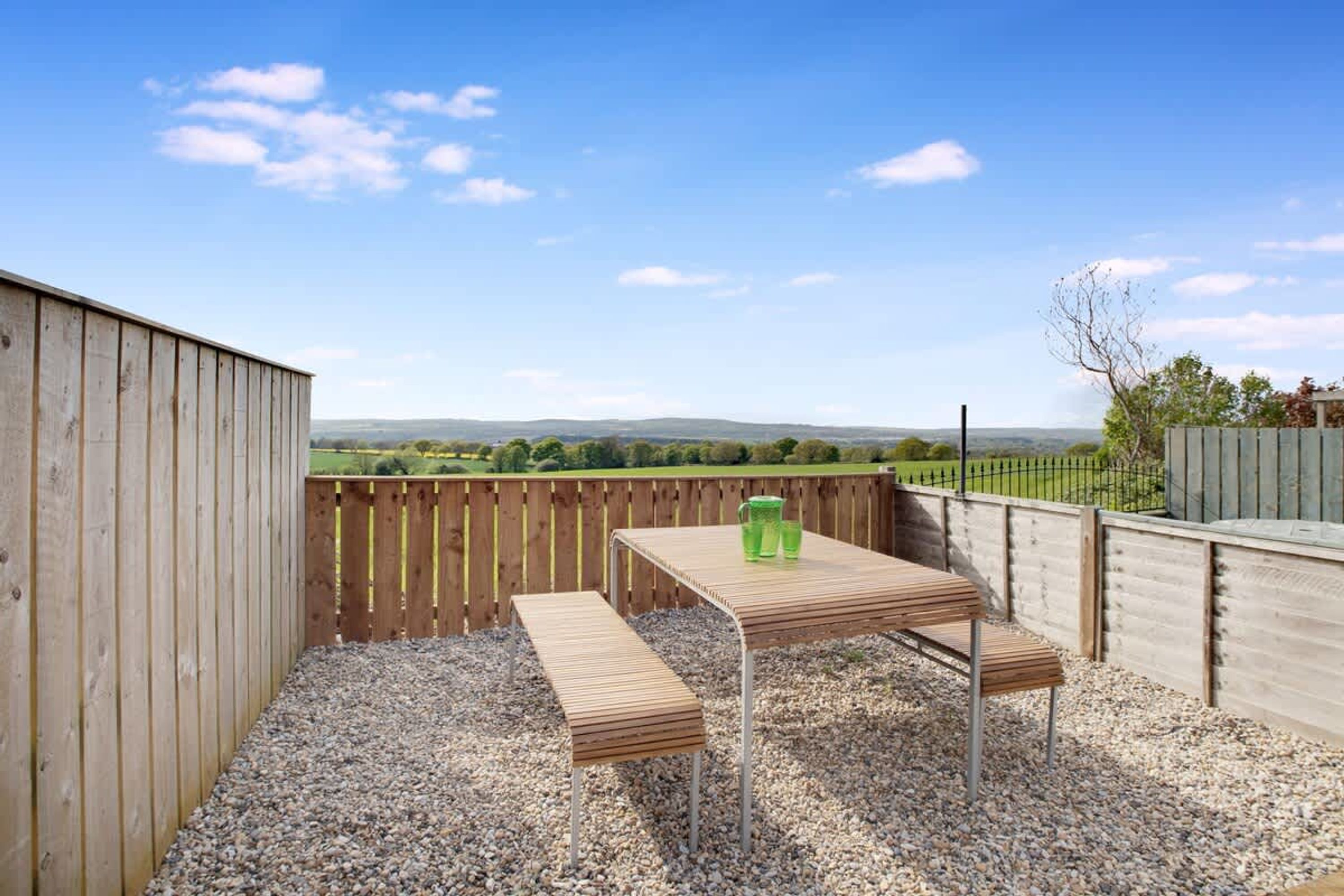 A wooden deck with a table and bench, gravel ground, and a view of a green landscape.