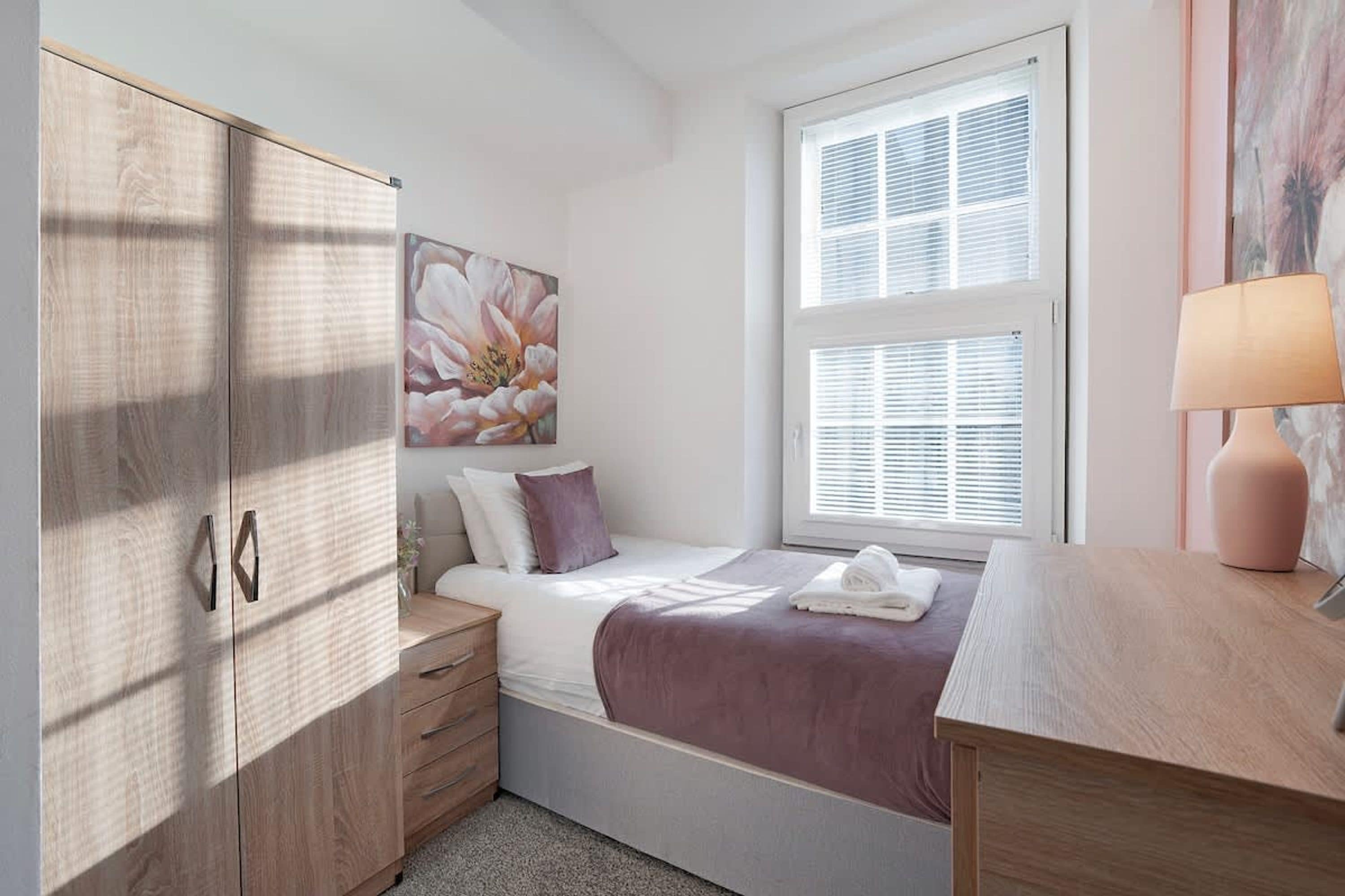 Ein Schlafzimmer mit Bett, Nachttisch, Schreibtisch, Lampe und einem Fenster mit Jalousien.