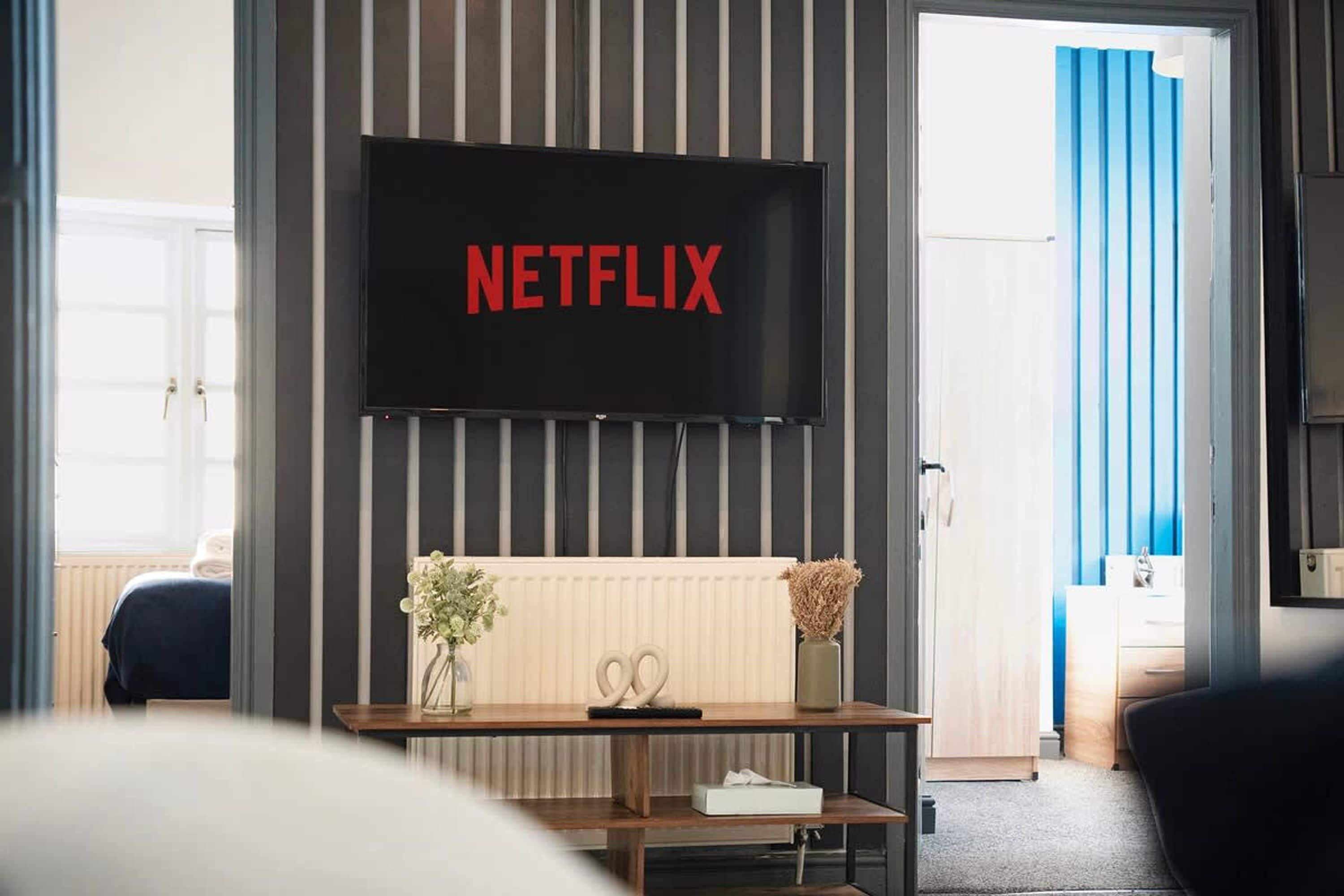 A room with a television mounted on the wall displaying 'NETFLIX' in red letters, a wooden console table with a vase and scissors, and a view of a blue curtain and a door.