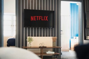 A room with a television mounted on the wall displaying 'NETFLIX' in red letters, a wooden console table with a vase and scissors, and a view of a blue curtain and a door. - 4 Bedroom With Private Parking and Wifi (Stockton-on-Tees)