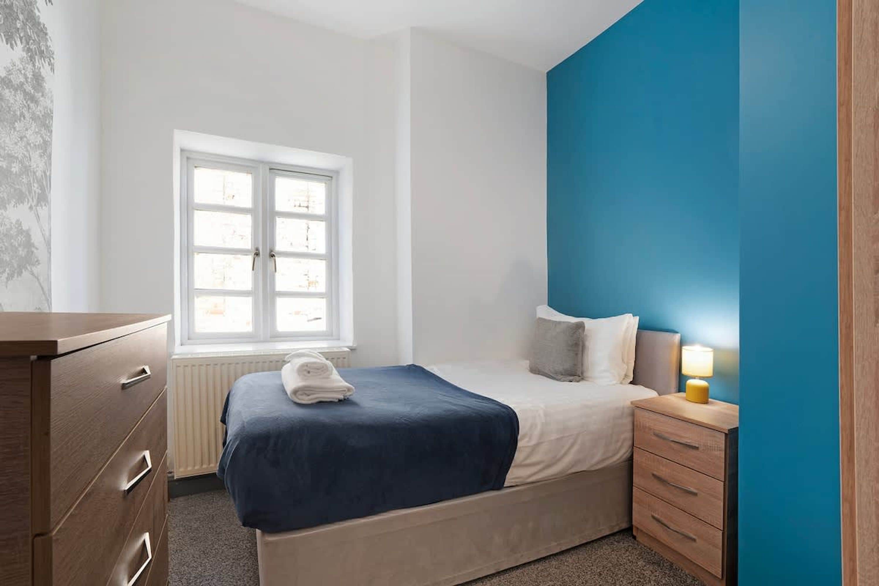 Un dormitorio con cama, mesita de noche, cómoda, ventana y una pared de acento azul.