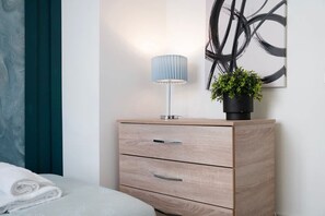 Een net opgemaakt bed met een gevouwen witte handdoek, een houten nachtkastje met een lamp en een potplant, en een muur met een groot abstract schilderij.