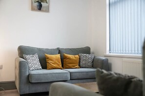 A grey sofa with patterned cushions, a potted plant on the wall, and a window with blinds. - Newcastle 2BR Sleeps 5 2 Parking by Stay With Us (Newcastle-upon-Tyne)