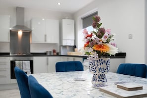 A modern kitchen with white cabinets, a stainless steel range hood, and a dining area with a blue chair and a vase of flowers on a marble table. - Newcastle 2BR Sleeps 5 2 Parking by Stay With Us (Newcastle-upon-Tyne)