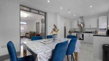 Una cocina moderna con gabinetes blancos, extractor de acero inoxidable y un comedor con mesa de mármol y sillas azules.