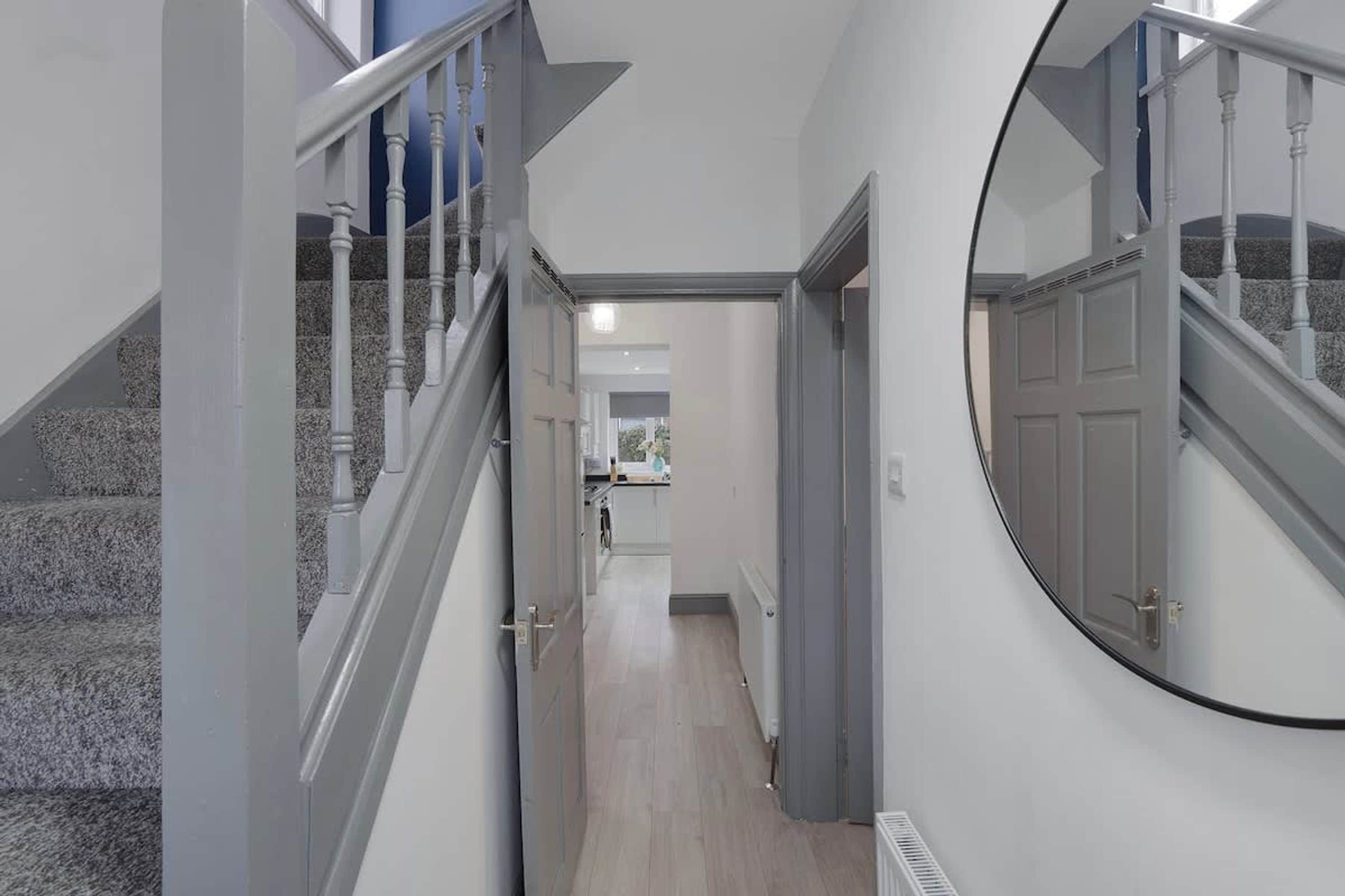 A hallway with a staircase on the left, a round mirror on the right, and a kitchen area in the background.