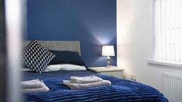 A bed with a blue blanket and pillows, a bedside lamp, and a window with blinds.