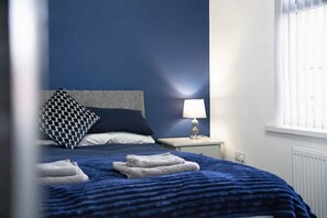 A bed with a blue blanket and pillows, a bedside lamp, and a window with blinds. - Newcastle 2BR Sleeps 5 2 Parking by Stay With Us (Newcastle-upon-Tyne)