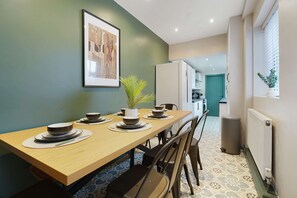 Un coin repas avec une longue table en bois dressée pour quatre personnes, un tableau encadré au mur et une plante en pot.