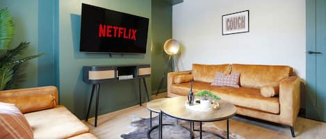 A living room with a sofa, a coffee table, a TV displaying the Netflix logo, and a framed sign that reads 'The Couch Club'.