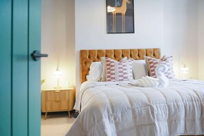 Un lit bien fait, avec une couette blanche et plusieurs oreillers, une table de chevet en bois avec une lampe, et une œuvre d’art représentant un cheval accrochée au mur.