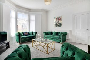 Una sala de estar con dos sofás verdes, una mesa de centro de cristal, un televisor y un cuadro en la pared.