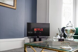 Una sala de estar con un televisor mostrando el logotipo de Netflix, una mesa de centro de vidrio y un sofá verde.