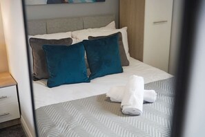 A neatly made bed with white linens and two teal pillows, a folded white towel on the bed, and a herringbone-patterned bedspread. - Modern 3 Bed Home With Garden Free Parking Wi-fi (Middlesbrough)