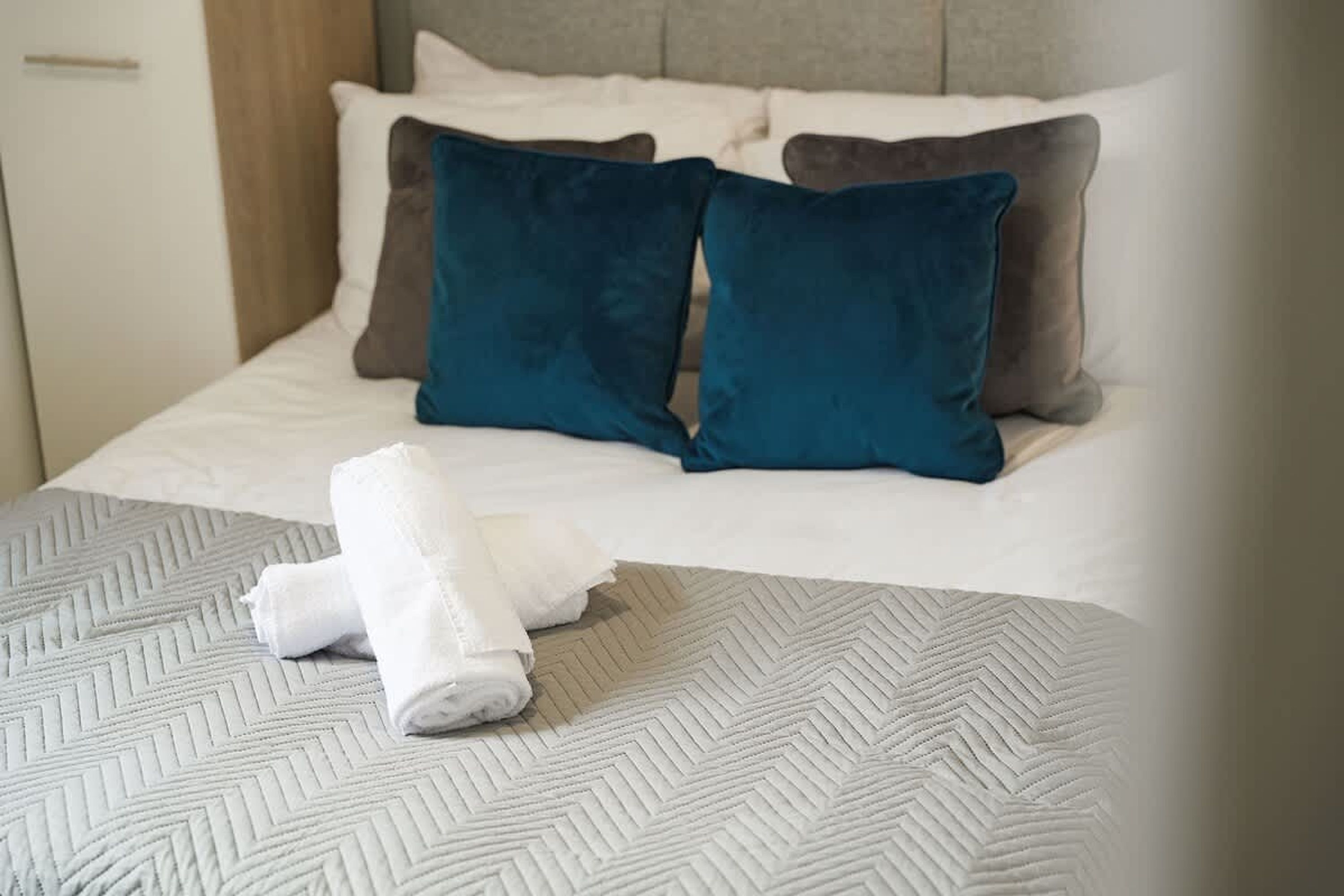 Een netjes opgemaakt bed met een beddekleed met patroon, twee kussens en een opgerolde handdoek aan de zijkant.