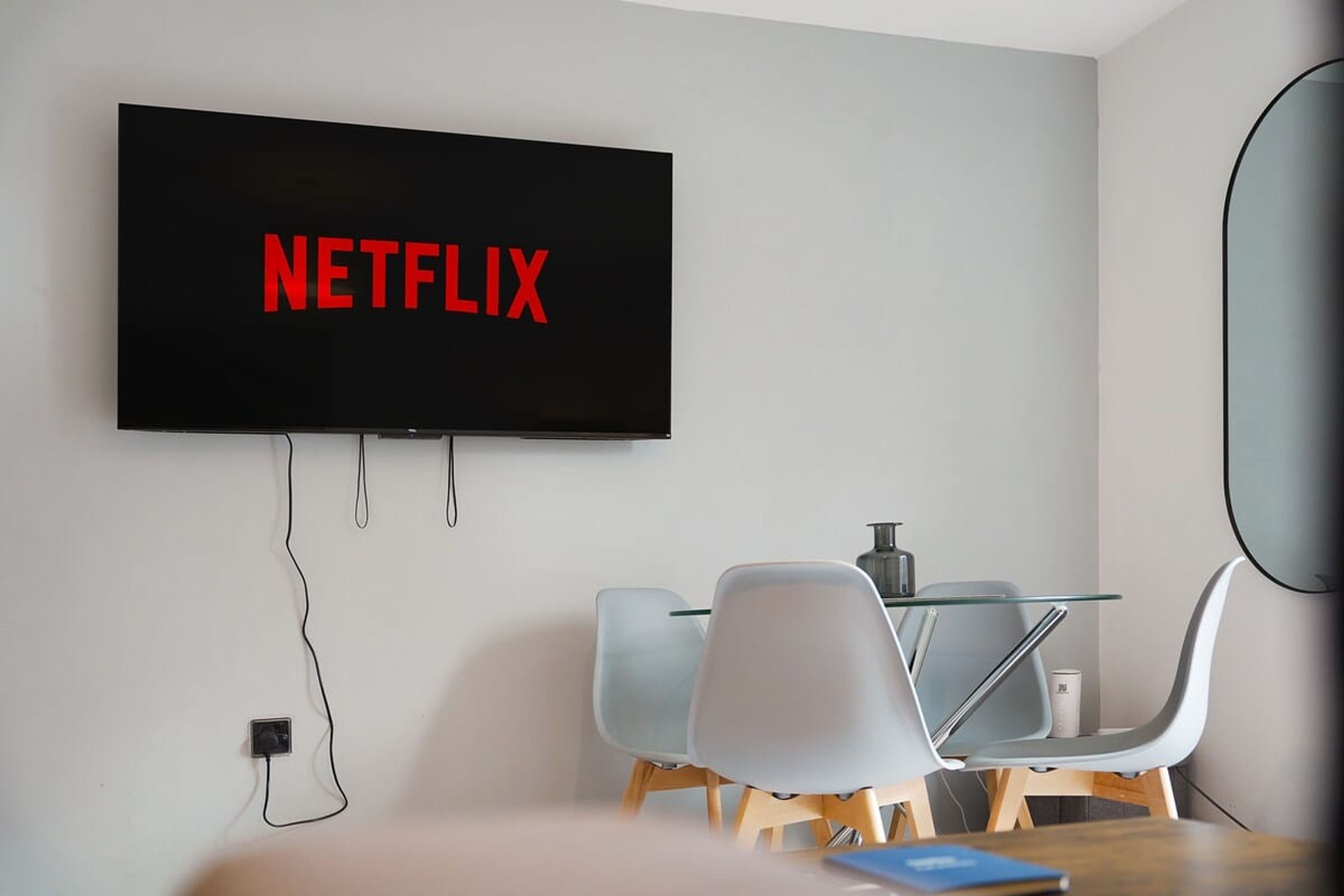 Ein modernes Zimmer mit einem wandmontierten Flachbildfernseher, der 'NETFLIX' in roten Buchstaben zeigt, einem Glastisch mit Stühlen und einer Steckdose an der Wand.