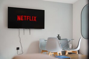 Ein modernes Zimmer mit einem wandmontierten Flachbildfernseher, der 'NETFLIX' in roten Buchstaben zeigt, einem Glastisch mit Stühlen und einer Steckdose an der Wand.