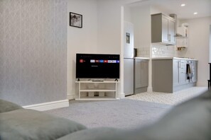 A living room with a flat-screen TV on a stand, a kitchen area with white cabinets, and a grey carpet. - Dean St Retreat Cosy Urban Escape Sleeps 5 (Hexham)