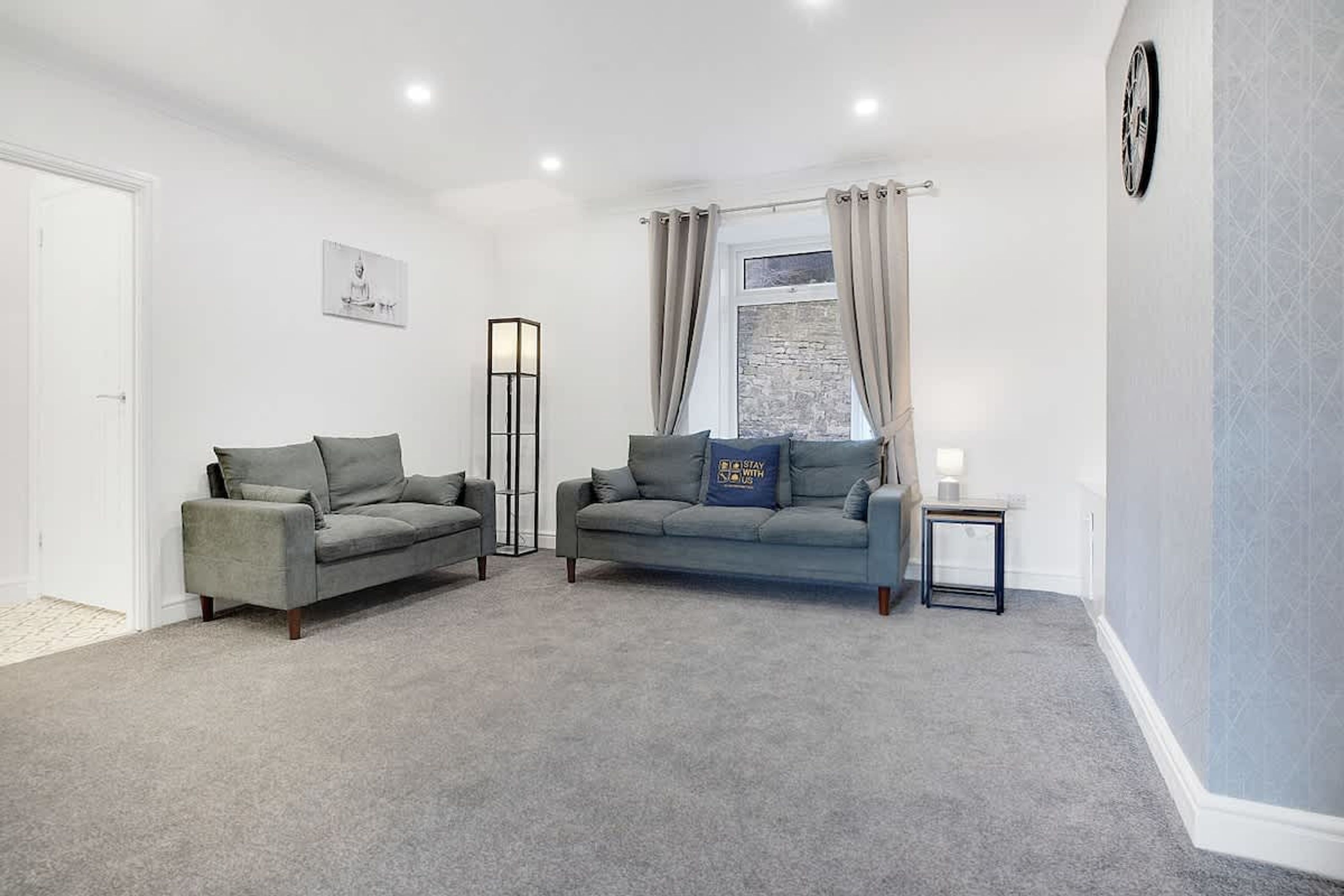 A living room with two grey sofas, a small table, a floor lamp, and a wall clock.
