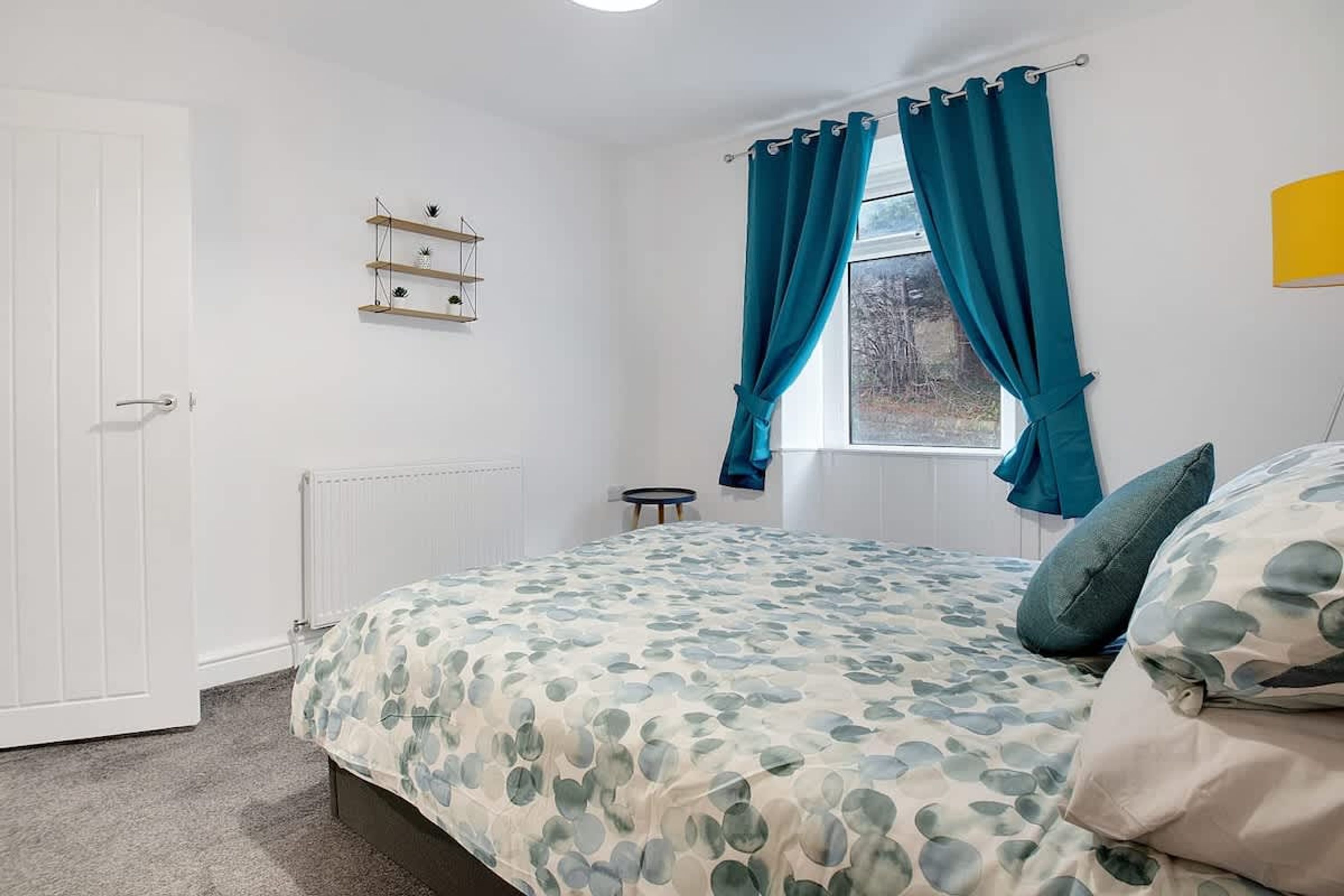 A bedroom with a bed, a window with curtains, a radiator, a small table, and a shelf with decorative items.