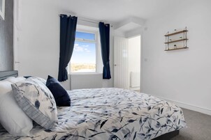 A bedroom with a bed, a window with curtains, a radiator, and a wall-mounted shelf with plants.