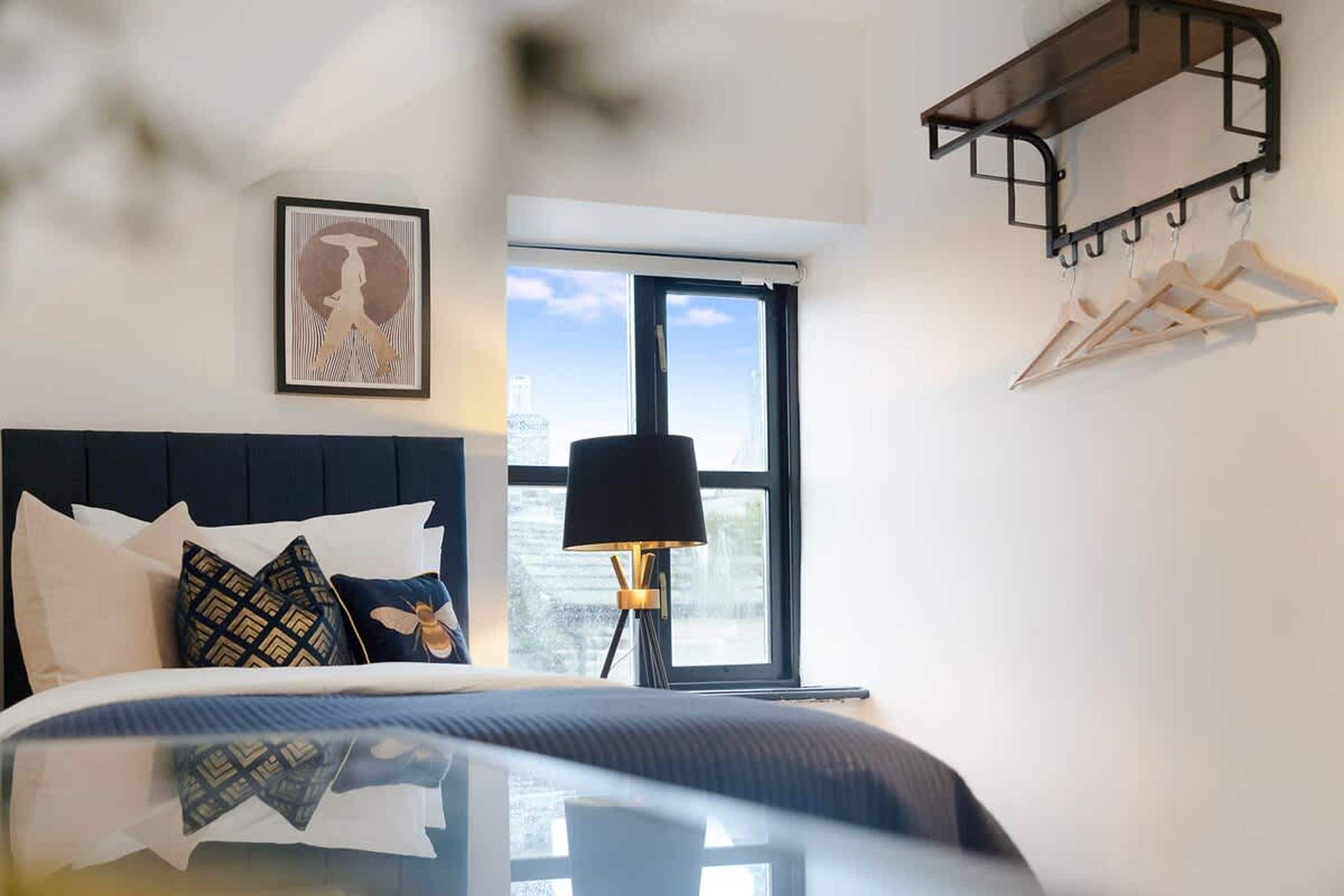 Quarto de hotel com cama, janela, luminária e cabideiro fixado na parede.