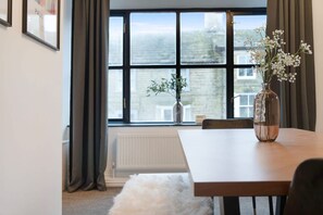 Une pièce avec une grande fenêtre, une table en bois, un vase rempli de fleurs et un tapis blanc et moelleux.