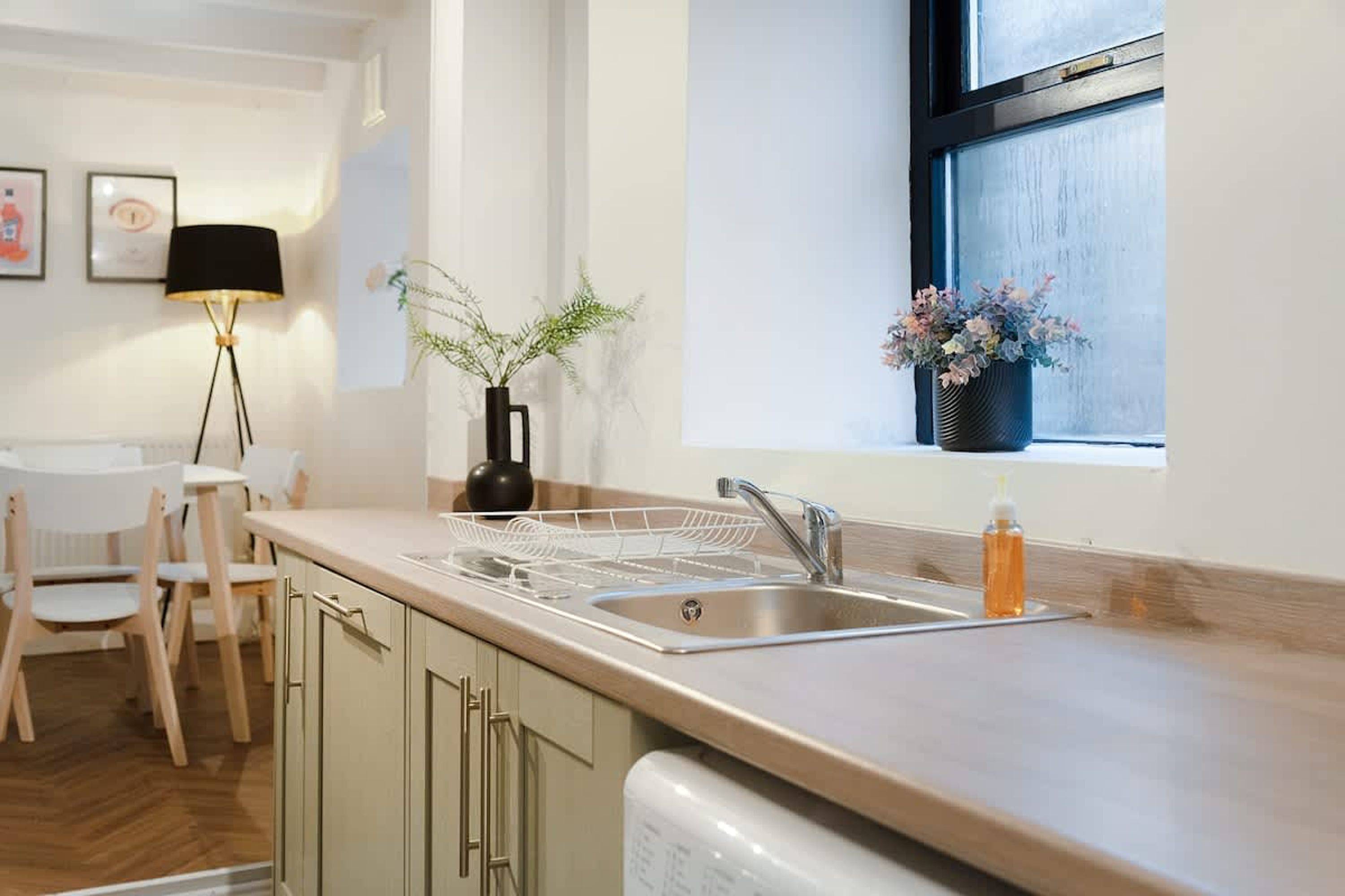 A modern kitchen with a stainless steel sink, wooden chairs, a black floor lamp, and a window with a view.