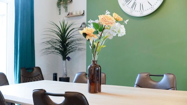 Un salón de conferencias con una mesa, sillas, un reloj y un florero con flores.