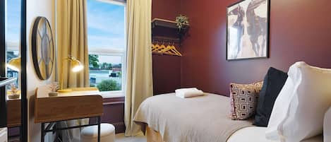 Habitación de hotel con cama, escritorio, espejo, ventana con cortinas, perchero y un cuadro en la pared.