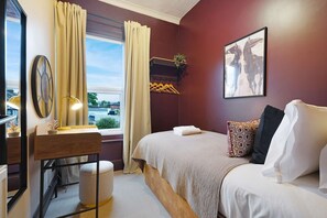 Une chambre d’hôtel comprenant un lit, un bureau, un miroir, une fenêtre avec des rideaux, un porte-manteau et un tableau encadré au mur.