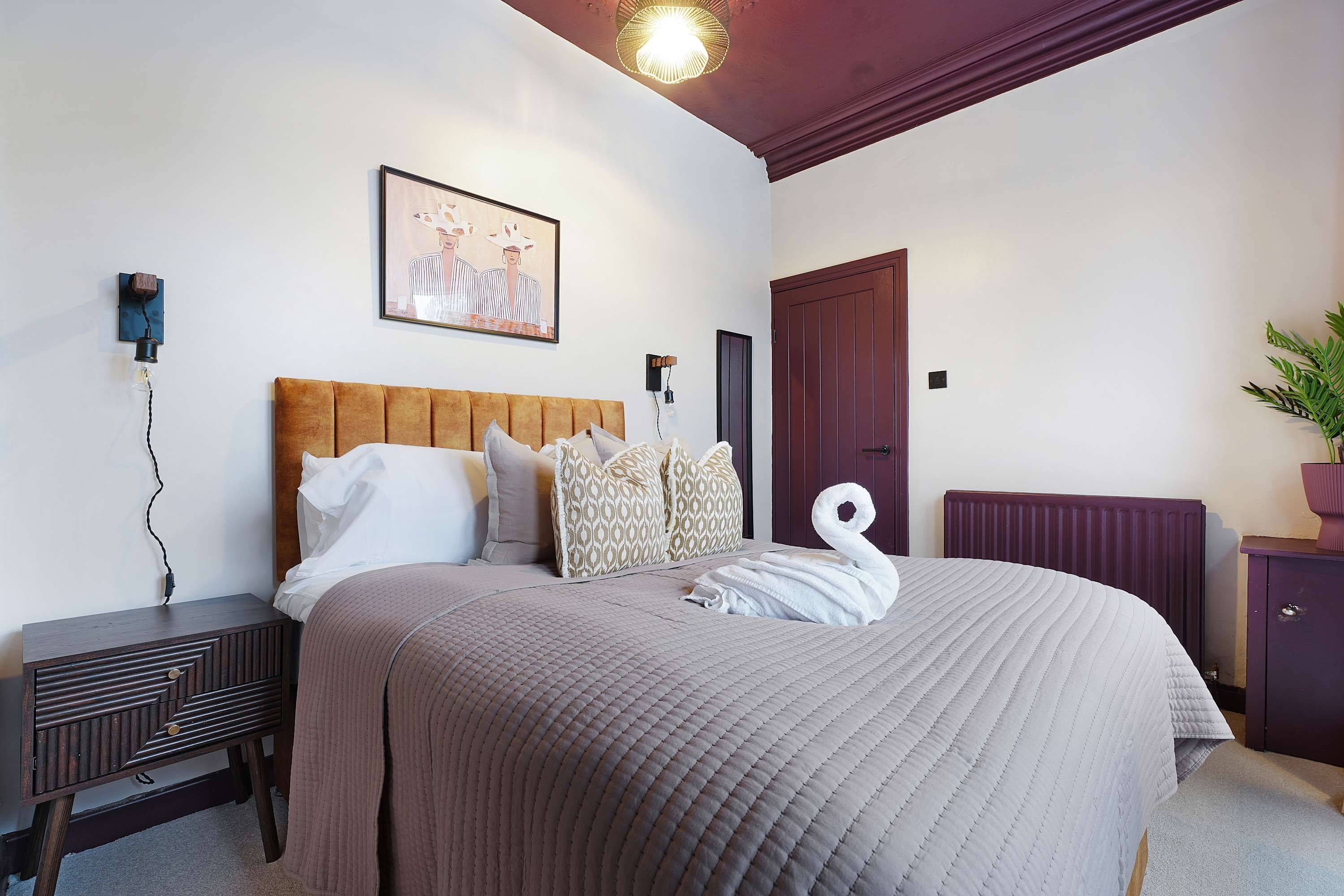 A bedroom with a bed, pillows, a headboard, a bedside table, a wall-mounted light, a door, and a radiator.