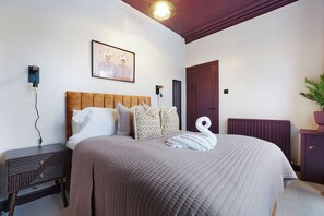 Ein Schlafzimmer mit Bett, Kissen, einem Kopfteil, einem Nachttisch, einer Wandleuchte, einer Tür und einem Heizkörper.