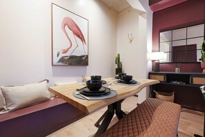 Ein Essbereich mit einem Holztisch, gedeckt für zwei Personen, einer Couch mit Kissen, einem Kaktus auf einem Regal und einem an der Wand hängenden Gemälde einer Flamingo.