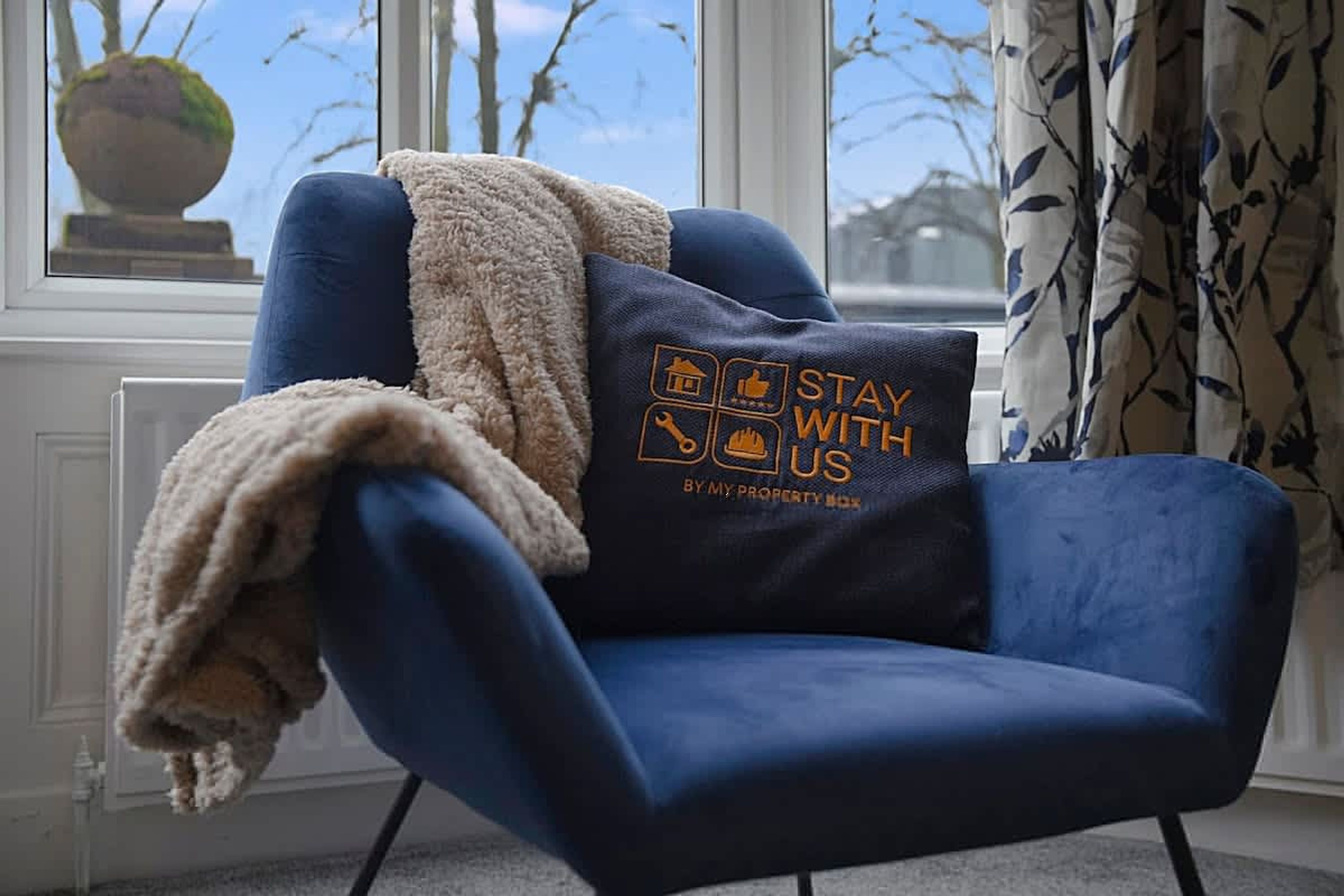 A blue armchair with a cushion featuring a stay with us logo, positioned by a window with a view of trees and a clear sky.