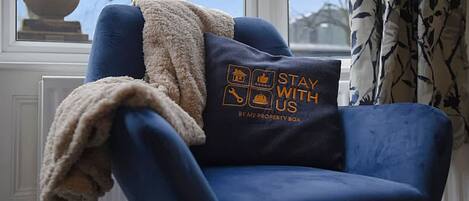 A blue armchair with a cushion featuring a stay with us logo, positioned by a window with a view of trees and a clear sky.