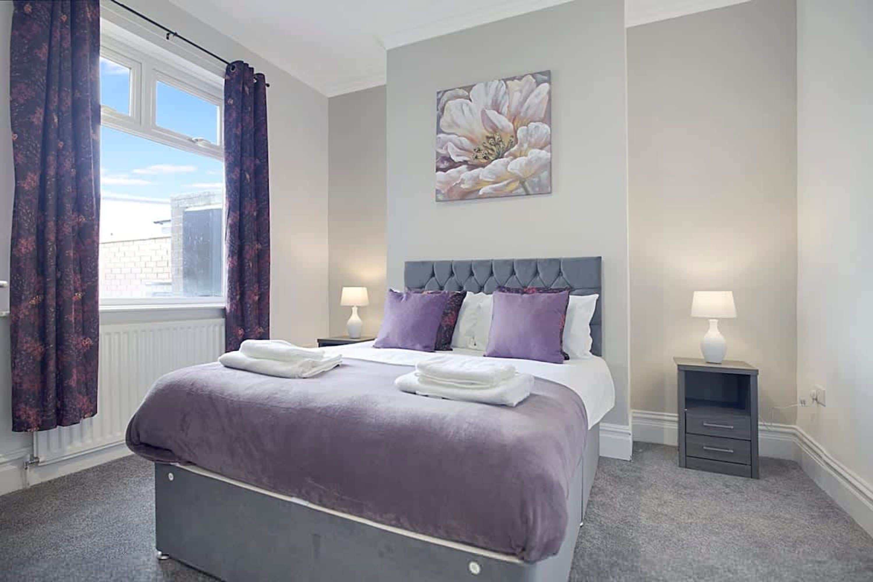 A hotel bedroom with a large bed, two bedside tables, a window with curtains, and a floral artwork on the wall.