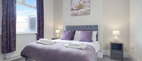 A hotel bedroom with a large bed, two bedside tables, a window with curtains, and a floral artwork on the wall.