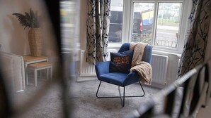 A room with a blue armchair, a small table, a potted plant, and a view of the outside through a window. - Spacious Retreat With Backyard Bliss Sleeps 7 (Bishop Auckland)