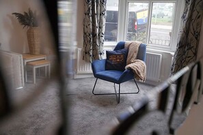 Ein Zimmer mit blauem Sessel, einem kleinen Tisch, einer Topfpflanze und Blick ins Freie durch ein Fenster.
