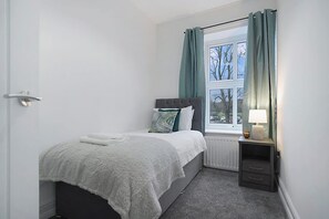 Une chambre à coucher avec un lit, une fenêtre avec des rideaux, une table de chevet avec une lampe et un radiateur.