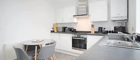 A modern kitchen with white cabinets, a built-in oven, a coffee machine, and a dining area with a round table and chairs.